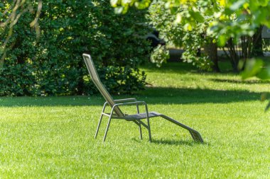 Çimenlikteki güverte sandalyesi. Tek bir iskemle, ağaçların gölgesinde güneşli bir çimenlikte duruyor. Burası sakin görünüyor ve sizi rahatlamaya davet ediyor..