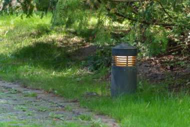 Yolun kenarındaki bahçe ışığı. Siyah bir bahçe ışığı, doğal taşlarla döşenmiş bir yolun kenarında parlıyor. Çimen ve ağaçlarla çevrili, sakin bir akşam atmosferi yaratır..