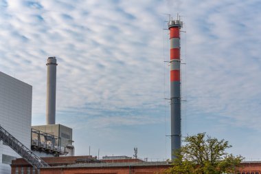 Kırmızı bantlı sanayi bacası. Kırmızı ve gri çizgili uzun bir endüstriyel baca bulutlu bir gökyüzüne yükselir. Çevreleyen fabrikada tuğla ve modern yapılar yer alıyor