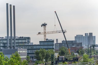 Bir vinç modern ofis ve sanayi binaları üzerinde bir patlama kolu kaldırır. Sahne, yeni binalar ve inşaat faaliyetleriyle kentsel değişimi resmediyor.