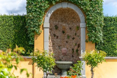 Garden 'daki duvar çeşmesi. Yüzü oyulmuş taş bir fıskiye suları yemyeşil bir havzaya püskürtür, yemyeşil bitkiler ve sarmaşıklarla çerçevelenir..