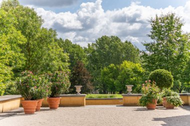 Çiçek açan zakkumlarla dolu, yemyeşil ağaçlarla çevrili güneşli bir teras. Sahne, parçalı bulutlu bir gökyüzünün altında sakin bir parka açılıyor..