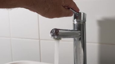 Musluk suyunu açıyorum. Bir el modern bir musluğu açar ve su akmaya başlar. Arka planda temiz beyaz karolar.