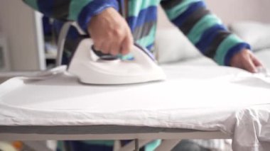 Bir el ütü masasında buharlı ütüyle beyaz bir elbise ütüler. Sahne, ütü sürecinin yakın çekimini yakalıyor..