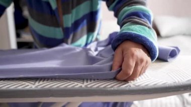 Mavi bir gömlek desenli bir tahta üzerinde ütülenir, elleri kumaşı gergin tutar. Buhar yavaşça yüzeyi yumuşatır..