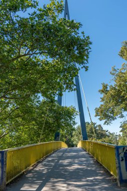 Sarı tırabzanlı yaya köprüsü. Sarı bir yaya köprüsü yeşil ağaçların arasından geçiyor. Yol çelik bir yapıya yükseliyor..