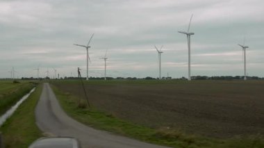 Manzarada rüzgar türbinleri var. Rüzgar türbinleri düz alanların üzerinde yükselirken, tren geçtikçe açık kırsal alanlarda yenilenebilir enerjiyi vurguluyor. Bir kanal ve yol mesafeye uzanıyor..
