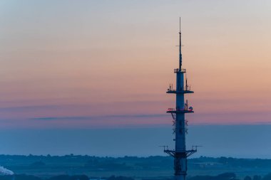 Pastel gökyüzüne doğru yükselen uzun bir iletim kulesi. Ufuk, yumuşak turuncu ve pembe renklerde uzanıyor..