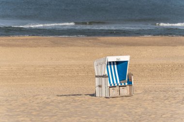 Mavi çizgili beyaz bir plaj sandalyesi kumlu sahilde tek başına duruyor. Sakin deniz ve dalgalar barışçıl bir zemin yaratır..