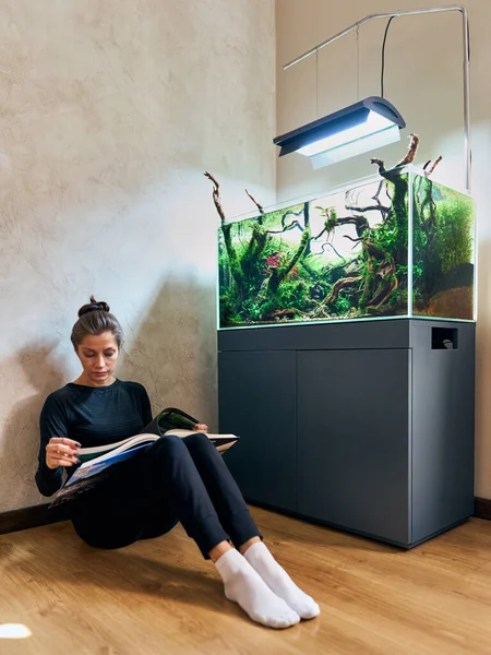 stock image A beautiful woman sitting on the floor and reading a book next to the aquarium