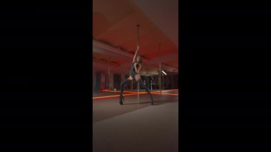 Yüksek topuklu uzun saçlı zayıf, esmer bir kadın kulüpte kırmızı ışıklı bir direğin üzerinde dans ediyor. Dikey 4K görüntü.