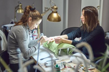 Happy beautician doing manicure to a smiling client under the gold lamp in the nail salon. Womans getting nail manicure.