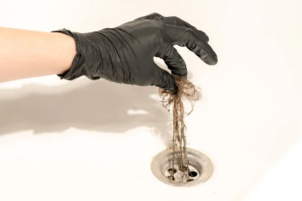 A bathroom drain clogged with dirty hair and slime. A female hand in a black latex glove pulls a long messy tuft of hair out of the drain