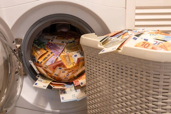 A big pile of money in an open washing machine. The big basket full of money is in front of. Money laundering concept.