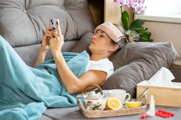 High temperature. A sick woman with a small towel on forehead lies under a blanket and holds a smartphone in her hands. In front of her there is a tray with a teapot, lemon and honey.