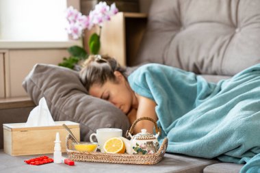 Grip salgını. Hasta bir kadın kanepede battaniyenin altında uyuyor. Önünde çaydanlık, limon ve bal dolu bir tepsi var. Yakınlarda haplar, termometre, burun spreyi ve dokular var. Bulanık yüz.