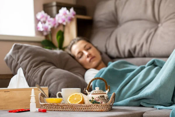 Flu epidemic. A sick woman sleeps under a blanket on the sofa. In front of her there is a tray with a teapot, lemon and honey. Nearby are pills, a thermometer, nasal spray and tissues. Blurry face.