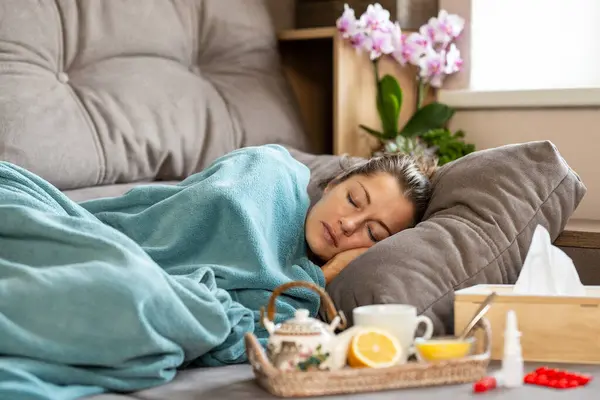 Flu epidemic. A sick woman sleeps under a blanket on the sofa. In front of her there is a tray with a teapot, lemon and honey. Nearby are pills, a thermometer, nasal spray and tissues.