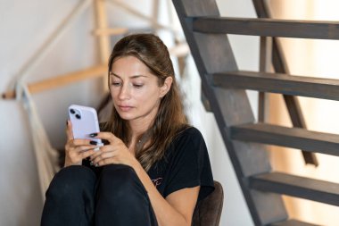 Evdeki ahşap merdivenlerin altında elinde telefonla oturan sevimli, gülümseyen genç kadın.