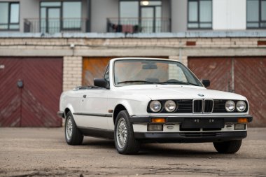 Riga, Letonya - 1 Nisan 2024: açık çatılı beyaz klasik üstü açık BMW E30 araba. Arka planda eski bir garaj ve modern bir bina..