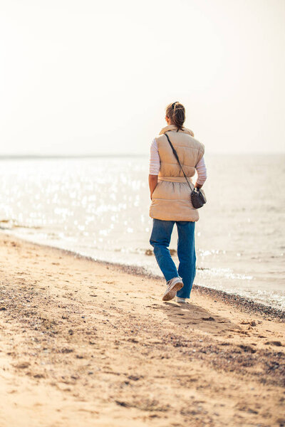Задний вид женщины, идущей вдоль пляжа, смотрящей на солнце. Вертикальный выстрел.