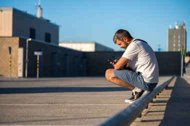 Olağanüstü düşünceli bir adam şehrin çatısında oturuyor ve güzel bir günbatımında elinde telefon arıyor.