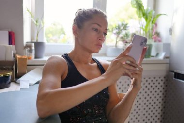 Parlak ve neşeli bir mutfak ortamında, kararlı genç bir kadın akıllı telefon cihazına odaklanır.