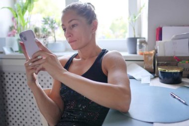 Parlak ve neşeli bir mutfak ortamında, kararlı genç bir kadın akıllı telefon cihazına odaklanır.