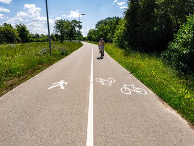 Bir bisikletçi, parlak mavi gökyüzünün altında yaya ve bisiklet sembolleriyle süslenmiş kaldırımlı bir yol boyunca mutlu bir şekilde süzülür.
