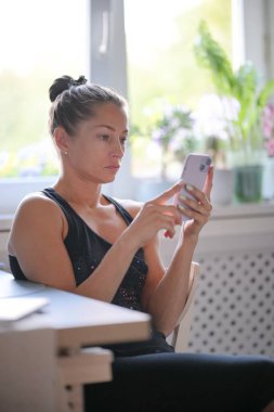 Parlak ve neşeli bir mutfak ortamında, kararlı genç bir kadın akıllı telefon cihazına odaklanır. Dikey