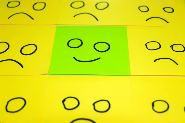 Unhappy and happy concept. Background of blue and green sticky notes. Happy sticky note is among unhappy sticky notes.
