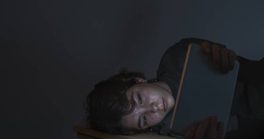 Genç bir kız loş bir odada elinde tabletle ya da elektronik aletle internette geziniyor.