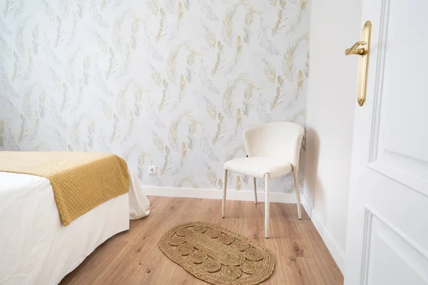 Bedroom with feathered wallpaper, white bed, yellow blanket, classic white chair, wooden floor and wicker rug. White door