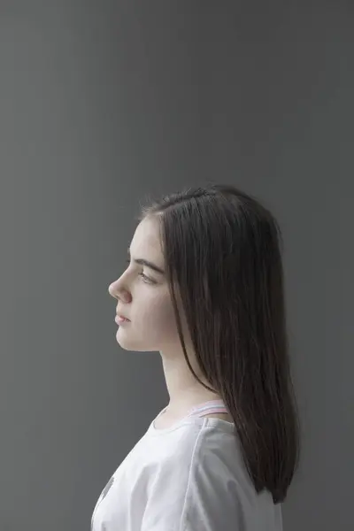 Vertical portrait with white background of a teenage girl in profile with long brown hair and a white t-shirt. With a serious expression
