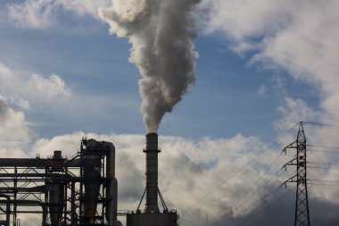 Ahşap işleme endüstrisinden çıkan dumanlı baca ve elektrik direği. Mavi gökyüzü ve beyaz bulutlu elektrik. Kirlilik