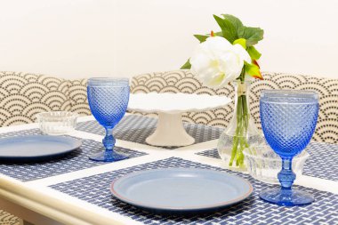 Mavi seramik tabaklar ve mavi saplı gözlüklerle bembeyaz ahşap mutfak masası. Beyaz ve mavi yaz dekorlu bir masa. Beyaz kamelyalı cam vazo.