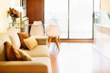 Bej renkli çift koltuklu oturma odası ve kahverengi yastıklar, doğal ışık ve beyaz sandalyeli cam masa.