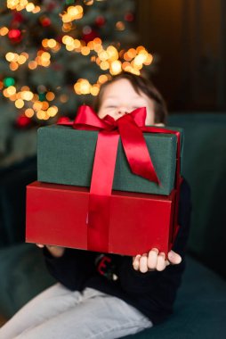 Çocuk Noel hediyelerini koltukta tutuyor. Arka planda, Noel sahnesinde.