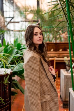 Uzun kahverengi saçlı, ceketli şık bir kadın serada, bitkilerin arasında.