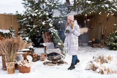 Hafif ceketli, elinde bir fincan çayla pembe şapkalı şık bir kız karlı bir yolda yürüyor.
