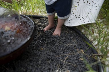 Küçük çocukların ayakları bahçedeki fıskiyenin yanındaki küçük siyah dekoratif taşların üzerinde duruyor.
