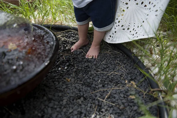 Küçük çocukların ayakları bahçedeki fıskiyenin yanındaki küçük siyah dekoratif taşların üzerinde duruyor.