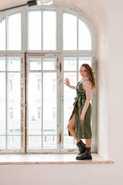 Uzun sarı kıvırcık saçlı, yeşil elbiseli ve dantelli ayakkabılı şık bir kız. Büyük, eski bir pencerenin penceresinde.