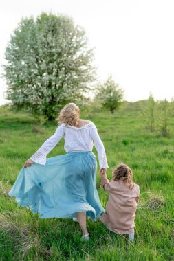 Genç, sarışın bir anne, küçük kızıyla çayırda yürüyor.