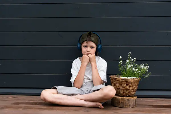 stock image Concentrated pre-teen boy in headphones sitting on a wooden terrace near a flower