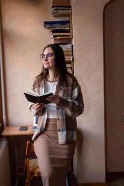 Gözlüklü serbest çalışan bir kız dizüstü bilgisayarlı bir masanın yanındaki defterde kalemle not alıyor.