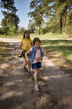 Okul çağındaki tatlı kızlar ormanda bir patikada jimnastik metal bir çemberle oynuyorlar.