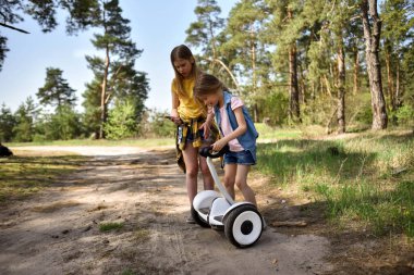 İki liseli kız, çam ormanında bir patikada scooter kullanıyor.