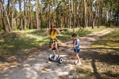 İki liseli kız, çam ormanında bir patikada scooter kullanıyor.