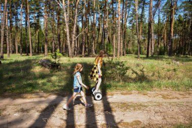 İki liseli kız, çam ormanında bir patikada scooter kullanıyor.
